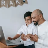 A father and son smiling while using a laptop, creating a moment of joy and bonding indoors.
